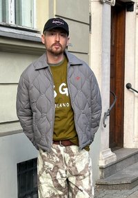 Casaco acolchoado cinza com colarinho largo, camisa verde com texto branco, calças de camuflagem com padrões castanhos e boné preto com logótipo.