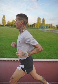 Persona tatuata con capelli corti che corre su una pista all'aperto, indossando una maglietta bianca, pantaloni neri, un orologio, una collana e anelli.