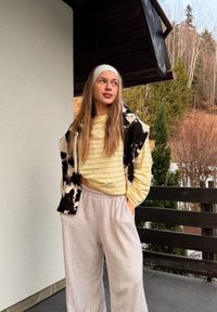 Νεαρή γυναίκα με κίτρινο ριγέ πουκάμισο και μπεζ παντελόνι στέκεται στη βεράντα με το χέρι στην τσέπη, κοιτάζοντας προς τα πλάγια. Διακρίνονται δέντρα και ξύλινο κιγκλίδωμα.