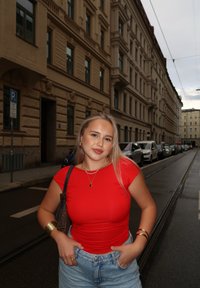 Rode T-shirt met korte mouwen, lichtblauwe high-waisted denim shorts, gelaagde gouden kettingen en armbanden; stedelijke straat met geparkeerde auto's op de achtergrond.