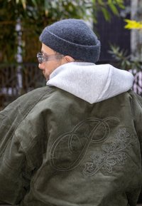 Veste en velours côtelé olive avec capuche de sweat-shirt grise, ornée d'un motif floral brodé et de la lettre "P" au dos, portée avec un bonnet gris et des lunettes.