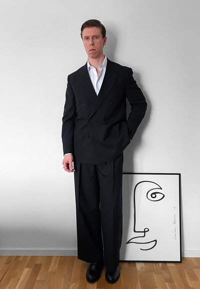 Hombre con traje negro de doble botonadura y camisa blanca, de pie contra una pared gris junto a un dibujo abstracto de una cara enmarcado sobre un suelo de madera.