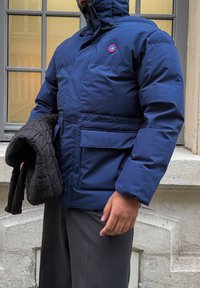 Chaqueta aislante azul marino con un cuello alto, cremallera y un emblema en el pecho. La persona sostiene un accesorio acolchado negro.