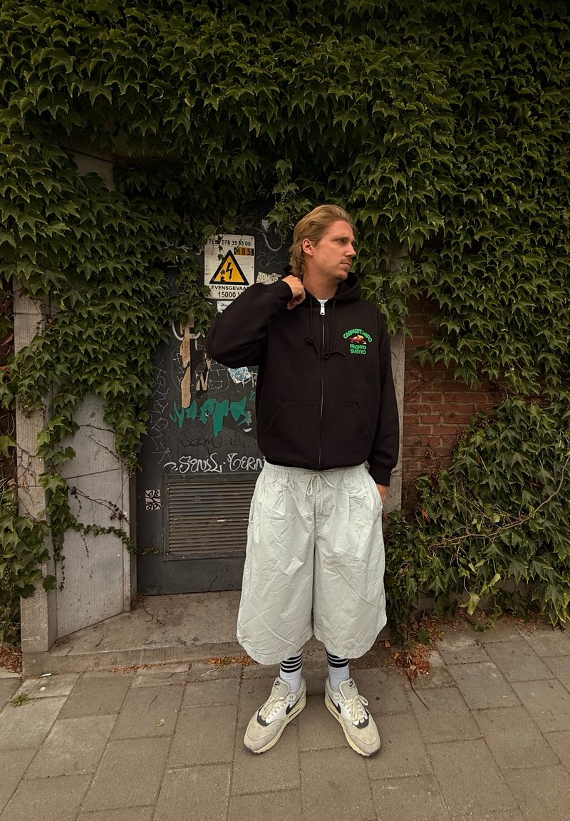 Sort hættetrøje med lynlås og grøn grafisk design, lysegrå oversized shorts og grå-hvide sneakers, stående foran en væg dækket af efeu.
