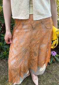 Lace skjørt i oransje med blomsterdesign, som dekker et lett stoff. Akkompagnert av en gul håndveske og beige sko.