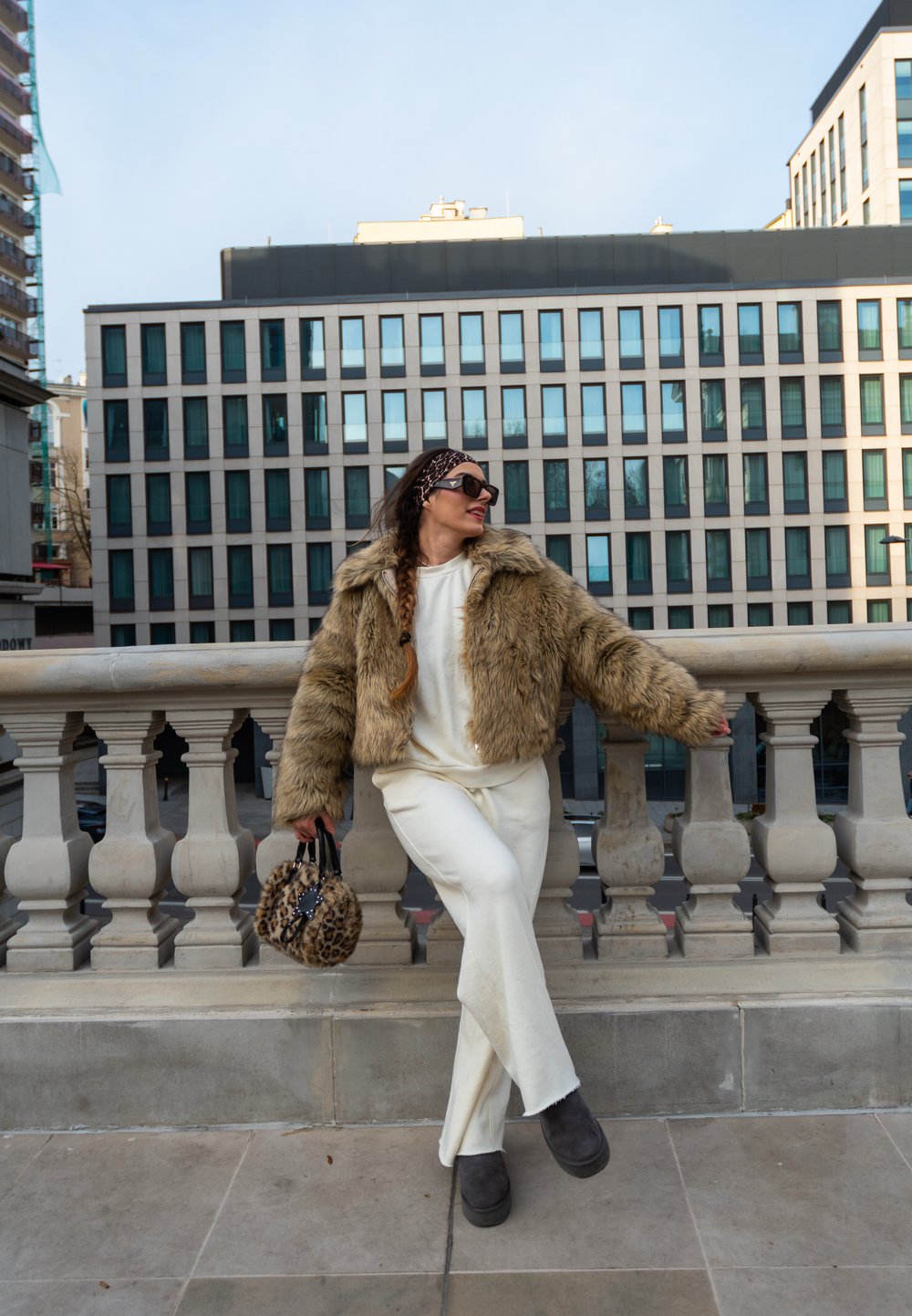 Brahmā mākslīgās kažokādas brūna jaka ar īsu piegriezumu; balta blūze un bikses; apaļa leopardu raksta rokas soma; lielas melnas saulesbrilles. Akmens margas fons.