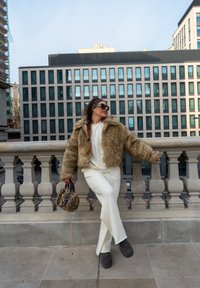 Brahmā mākslīgās kažokādas brūna jaka ar īsu piegriezumu; balta blūze un bikses; apaļa leopardu raksta rokas soma; lielas melnas saulesbrilles. Akmens margas fons.