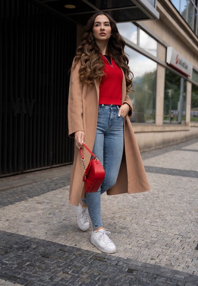 Abrigo largo beige, top rojo con cremallera, jeans de cintura alta azul claro, zapatillas blancas y un pequeño bolso rojo con correa. Entorno urbano.