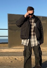 Doudoune noire portée sur une chemise à carreaux grise aux bords effilochés, associée à un pantalon cargo noir avec de multiples poches, sur fond de bord de mer.