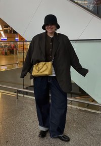 Oversized svart blazer over en brun knappebluse, kombinert med brede blå jeans. Gul håndveske med håndtak og svarte sko. Svart bøttehatt.