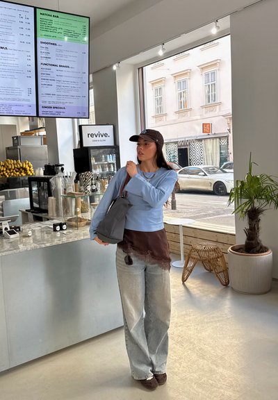 Mujer con ropa casual y gorra sosteniendo una bolsa dentro de un café luminoso cerca de un mostrador con plátanos y un menú digital en la parte superior.