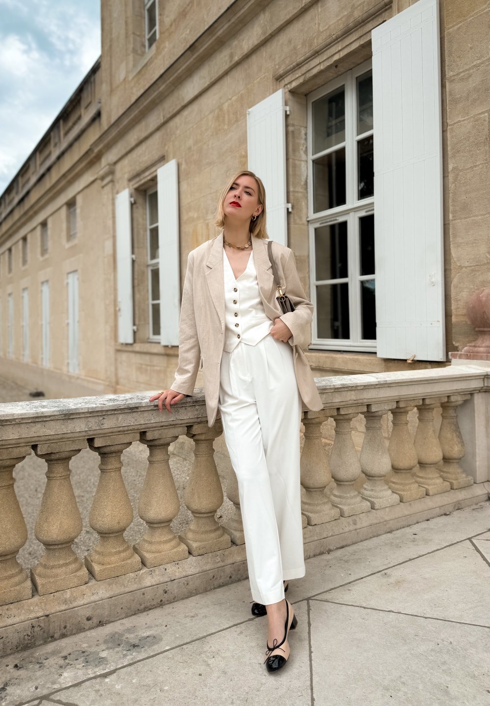 Lichtbeige linnen blazer over een wit overhemd en wijde broek, gecombineerd met zwartbeige flats, op de achtergrond van een stenen balustrade.