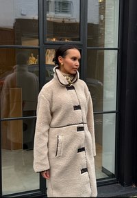 Bézs színű, műszőrme kabát magas gallérral, fekete gombos z�áródással és kontrasztos szegéllyel. Oldalzsebekkel és kényelmes tapintással rendelkezik.