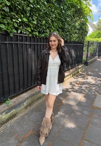 Casaco bomber de couro castanho sobre um vestido mini branco, combinado com botas de tornozelo com franjas bege; o fundo apresenta folhagem verde e uma cerca preta.