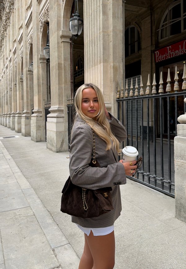 Jeune femme aux longs cheveux blonds, portant un blazer à carreaux et un short blanc, tenant une tasse de café et un sac près de colonnes en pierre classiques.