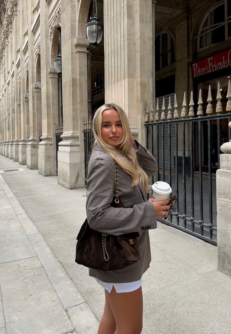 Jeune femme aux longs cheveux blonds, portant un blazer à carreaux et un short blanc, tenant une tasse de café et un sac près de colonnes en pierre classiques.