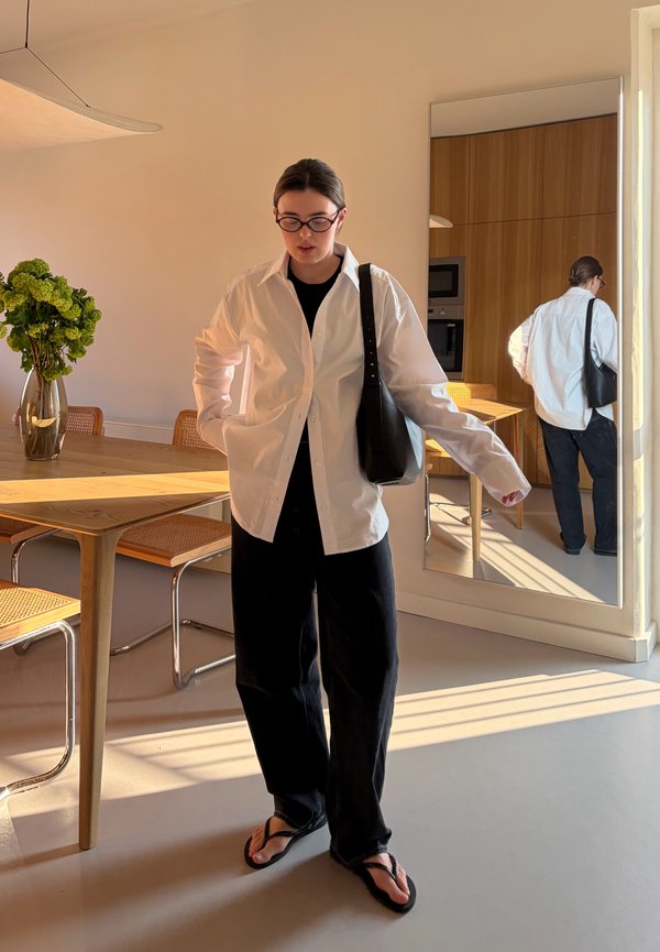 Donna con camicia bianca, pantaloni neri e infradito si trova in una stanza illuminata dal sole con un tavolo di legno, sedie e un grande specchio.