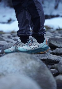 Chaussures de trail running en bleu clair et gris avec des détails réfléchissants, une tige en mesh et une semelle extérieure robuste avec des accents orange, conçues pour une utilisation en extérieur.