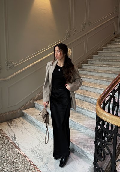 Blazer gris sobre una blusa negra y una falda larga negra; sosteniendo un pequeño bolso marrón con una correa. Escalera de mármol y barandilla ornamentada de fondo.