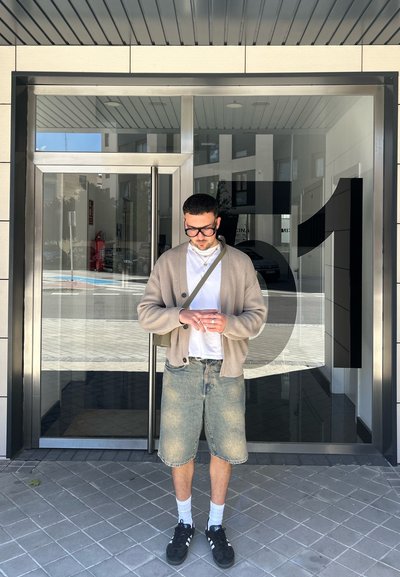 Cardigan beige claro sobre una camisa blanca, pantalones cortos de denim azul y zapatos negros con calcetines blancos, de pie frente a una puerta de vidrio marcada con el número '51'.