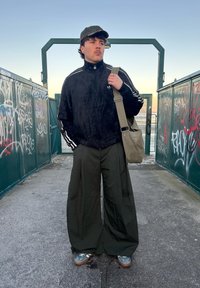 Black fuzzy jacket with white stripes, olive green wide-leg trousers, grey trainers, and a dark cap, holding a light olive bag. Graffiti in the background.