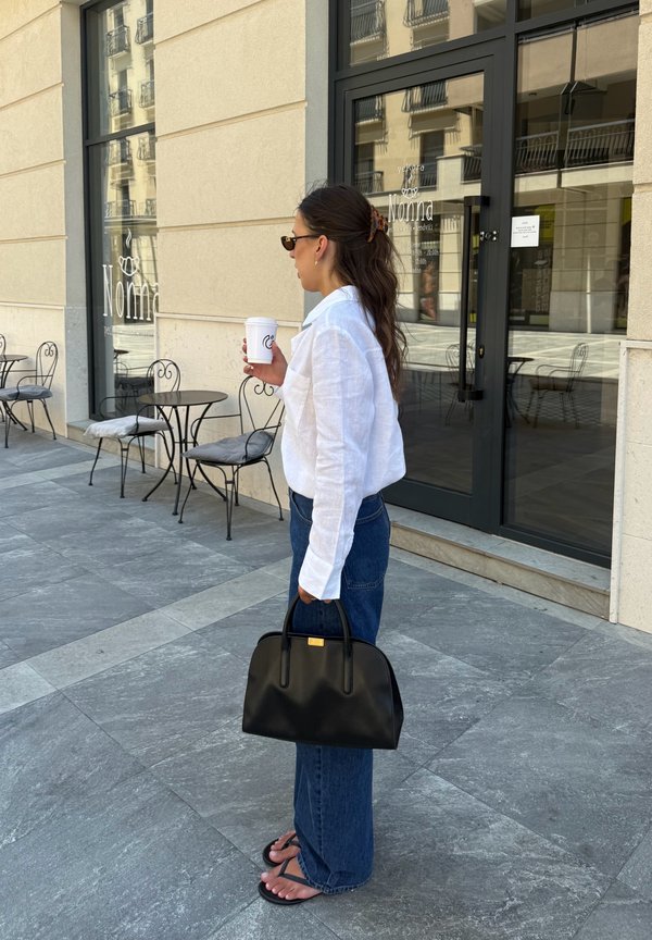 Donna in camicetta bianca e jeans che tiene una borsa nera e una tazza di caffè, in piedi fuori da un caffè con tavoli e sedie in metallo.