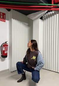 Femme accroupie dans un parking, regardant un extincteur rouge fixé sur un mur blanc ondulé avec des tuyaux rouges et verts apparents au-dessus.