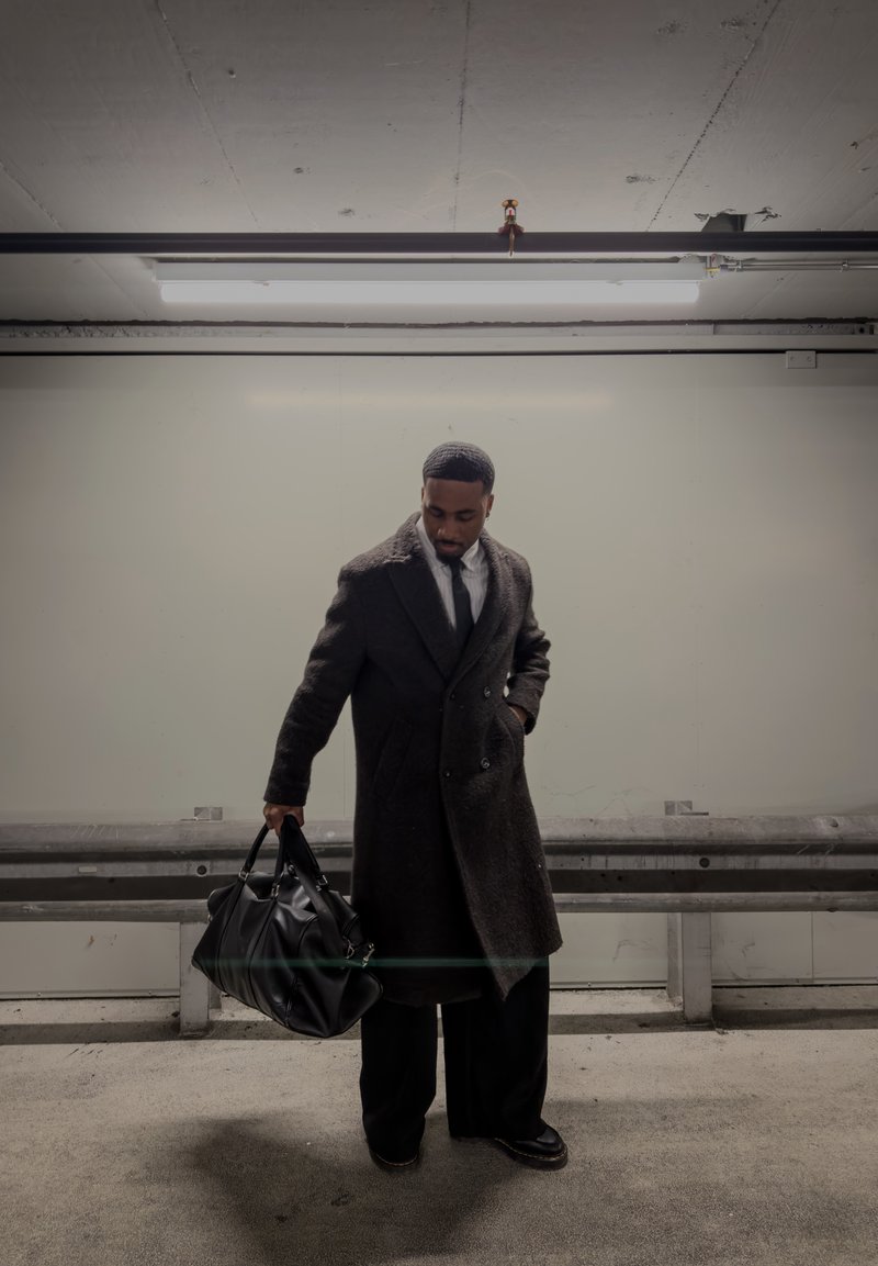 Homme portant un long manteau sombre et une cravate se tient dans un parking faiblement éclairé, tenant un grand sac de voyage en cuir noir dans sa main gauche.
