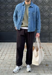 Jeans jakna v svetlo modri barvi, olivno zelena pulover pod njo, črne hlače in bele superge, drži bež torbo. Tekstura ozadja.