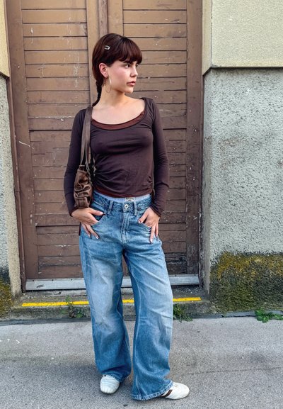 Top de manga larga marrón, jeans anchos de color azul claro, zapatillas blancas. Accesorio: pequeño bolso de hombro marrón. Fondo: puerta de madera y pared de hormigón.