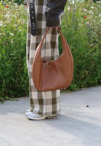 Καφέ δερμάτινη τσάντα ώμου με στρογγυλή μορφή, αντίθετη λευκή ραφή και κλείσιμο με φερμουάρ που διαθέτει γεωμετρικό διακοσμητικό στοιχείο.