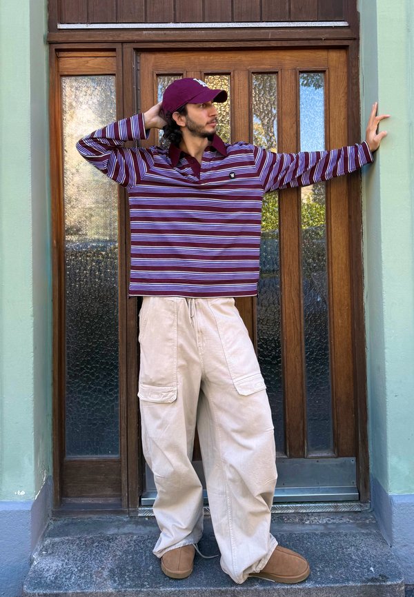 Giovane uomo che indossa una camicia a righe e pantaloni cargo beige, in piedi con una mano sulla testa e l'altra sul telaio di una porta in legno.