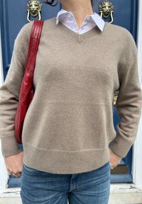 Béžový pletený sveter s výstrihom do V, textúrovaná látka a rebrované manžety, kombinovaný s červenou koženou ramennou taškou pred modrými dverami.