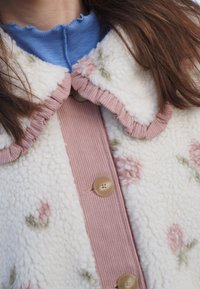 Veste en polaire blanche avec motif floral rose, dotée d'une patte de boutonnage et d'un col côtelés roses aux bords froncés, agrémentée de boutons ronds.