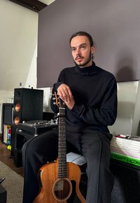 Guitarra acústica com corpo de madeira, acabamento natural e braço preto, segurada por uma pessoa vestindo um suéter de gola alta preto, sentada perto de equipamento de áudio.
