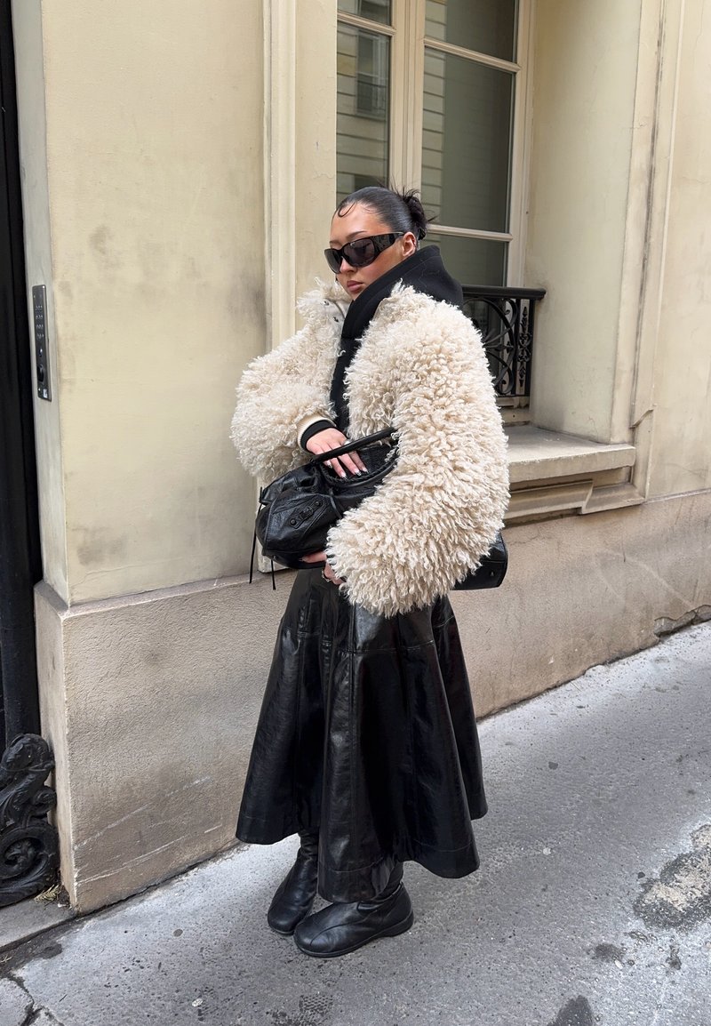 Femme portant une veste en fausse fourrure blanche surdimensionnée, une jupe en cuir noir, des bottes noires, des lunettes de soleil, et tenant un sac à main noir dans une rue de la ville.