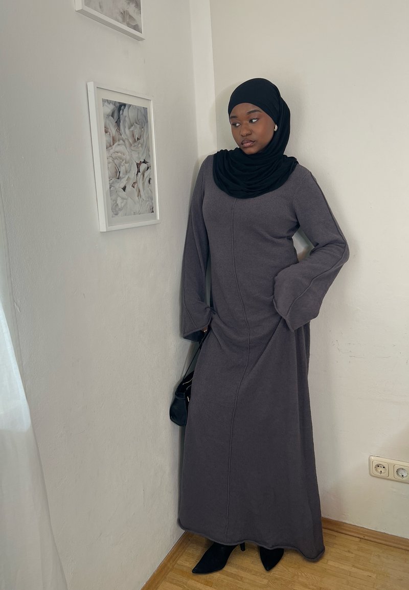 Sieviete melnā hijabā un garā pelēkā kleitā stāv pie baltas sienas netālu no iestiklotas baltas ziedu mākslas un koka grīdas.