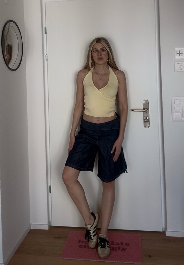 Giovane donna con occhiali che indossa un top giallo halter, pantaloncini scuri sopra il ginocchio e sneakers, in piedi davanti a una porta bianca chiusa all'interno.