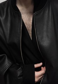 Veste aviateur en cuir noir avec fermeture éclair frontale, poignets côtelés et texture lisse, portée par-dessus un vêtement noir à encolure en V profonde.