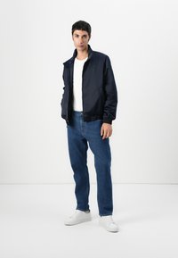 Tengeri bomber dzseki felálló gallérral, kék farmernadrággal és fehér tornacipővel párosítva. Sima anyag, rugalmas mandzsettákkal és derékpánttal.