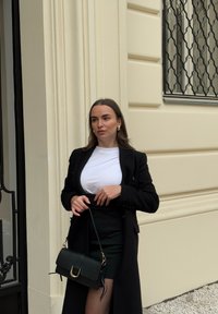Cappotto nero su una maglietta bianca e gonna nera. Tiene una piccola borsa nera con accessori dorati. Texture lisce e colori neutri.