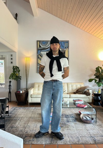 Camiseta blanca, suéter negro atado alrededor del cuello, vaqueros sueltos de color azul claro, zapatillas negras. Ambiente interior con un sofá de color claro y vegetación.