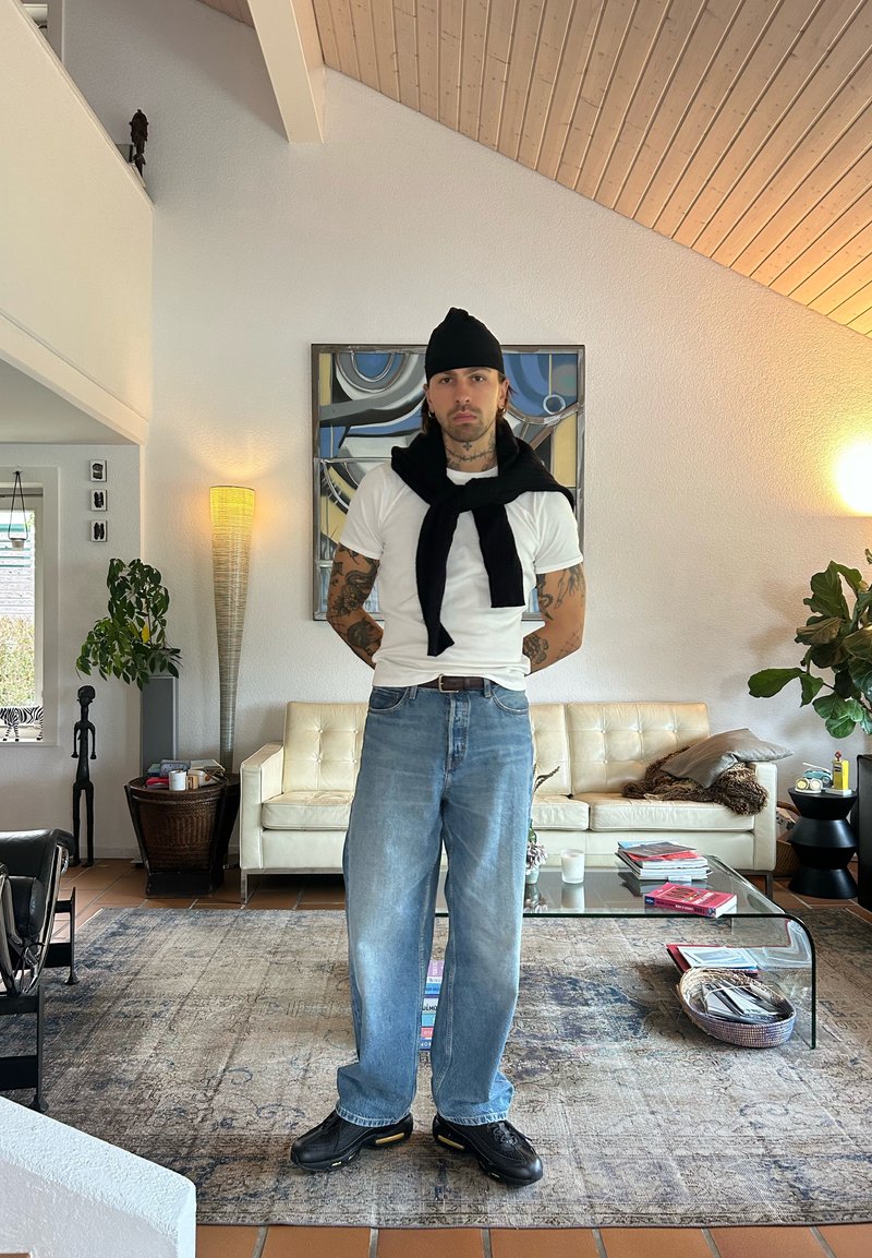 Witte t-shirt, zwarte trui om de nek geknoopt, lichtblauwe losse jeans, zwarte sneakers. Binnenomgeving met een lichtgekleurde bank en groenvoorzieningen.