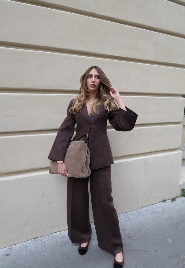 Ung kvinde med langt hår iført en brun buksedragt og holder en stor beige håndtaske, står op ad en beige væg udendørs.