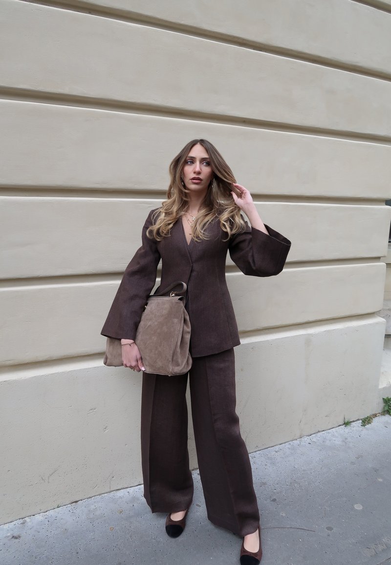 Νεαρή γυναίκα με μακριά μαλλιά, φορώντας καφέ κοστούμι με παντελόνι και κρατώντας μια μεγάλη μπεζ τσάντα, στέκεται μπροστά σε έναν μπεζ τοίχο έξω.