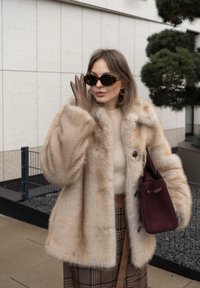 Borsa in pelle scamosciata bordeaux, cappotto in pelliccia beige con texture morbida, maglione beige a costine, gonna a quadri e occhiali da sole tartarugati.