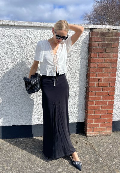 Blusa blanca corta con borde festoneado, atada en el frente, combinada con una falda maxi negra. Accesorios incluyen un bolso negro texturizado y tacones.