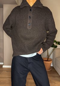 Pull marron côtelé avec un col à revers et trois boutons noirs. Porté avec un pantalon foncé, mettant en évidence un sous-pull blanc visible.