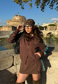 Bruine jas met ritssluiting met turquoise strepen, bijpassende bruine rok en kniehoge bruine laarzen. Afgewerkt met een hoofddoek met luipaardprint en een bril.