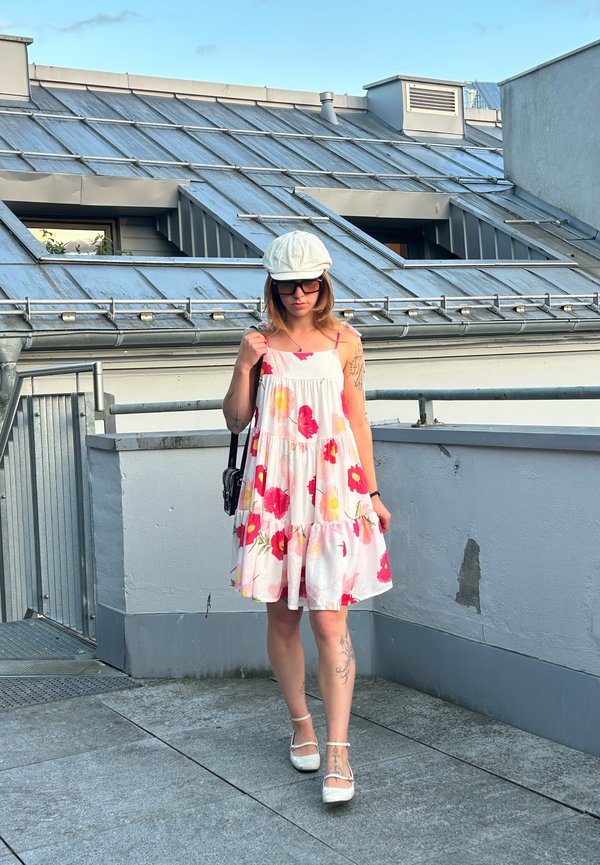 Jeune femme tatouée portant une robe blanche à fleurs, un chapeau blanc, des lunettes de soleil et des chaussures blanches, debout sur une terrasse sur le toit avec une rambarde en métal.